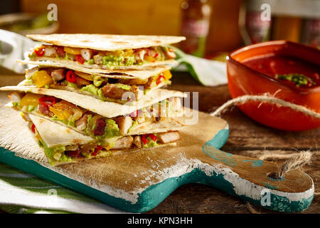 Vecchio tagliere in legno con servizio di tex-mex Turchia o di maiale quesadilla antipasti anche riempito con il guacamole, peperoni e pomodori con salsa ciotola in background Foto Stock