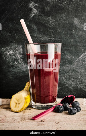Singolo bicchiere di deliziosi Acai berry smoothie tra una fetta di banana, mirtilli e cucchiaio di plastica su Sfondo marmo Foto Stock