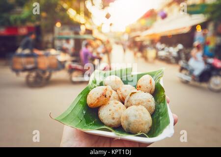 Mano di uomo tenendo la coppa con noce di cocco dolci cibo. Strada trafficata piena di ristoranti, bar e negozi - Siem Reap, Cambogia Foto Stock