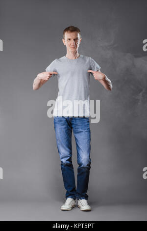 La mezza età uomo in jeans dita di puntamento per la sua t-shirt su sfondo grigio. Foto Stock
