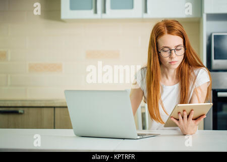 Redhead utilizzando il tablet PC e notebook in cucina Foto Stock