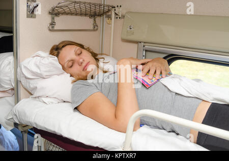 La ragazza in un treno dorme sul retro del ripiano superiore e il lato con la rivista in un abbraccio Foto Stock