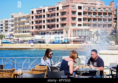 Famiglia seduti a un tavolo ristorante lungo il mare con la spiaggia e la città verso la parte posteriore e le onde si infrangono contro la parete promenade, Marsalfor Foto Stock