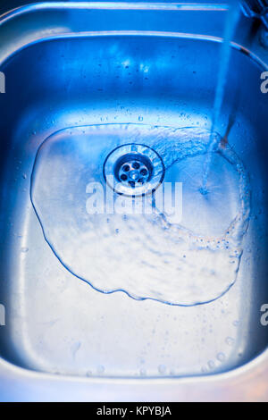 Un flusso di pulito e trasparente di acqua fresca fluisce dall'acciaio lucido toccare in un lavello in acciaio inox nei toni del blu. Scarichi acqua nel foro per TH Foto Stock