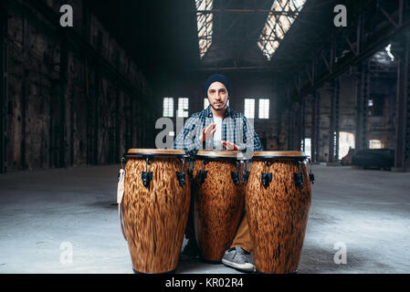 Batterista maschio suona sul tamburo di legno nel Grande negozio della fabbrica, vista frontale, musicista in movimento. Bongo, musicale strumento a percussione, musica etnica Foto Stock