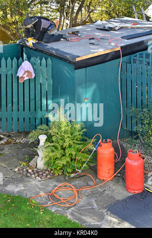 Copritetti lavorando sulla re-copertura piana che perde garden shed premendo verso il basso sul bitume riscaldato adesivo sul nuovo bitumati propano di bombole di gas al di sotto di Foto Stock