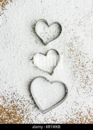 Tre a forma di cuore ad tagliapasta su una pasta ricoperta di zucchero a velo. Queste frese sono usati per rendere tradizionali biscotti di Natale. Amore per cuocere il concetto Foto Stock