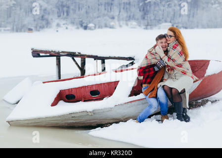 A tutta lunghezza shot della bella amare giovane avvolto in maglia plaid e seduta sulla barca vicino al fiume congelato durante la nevicata. Foto Stock