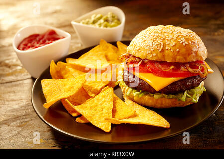 Deliziosa grandi cheeseburger e rabboccato con pomodori e cipolle a fianco triangolare di nacho chips e ciotole di salsa guacamole e Foto Stock