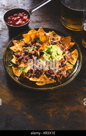 Salsa rossa salsa calda nella ciotola accanto al grande piastra in ceramica di mais giallo tortilla chips, guacamole e birra scura sul tavolo di legno con lo spazio di copia Foto Stock