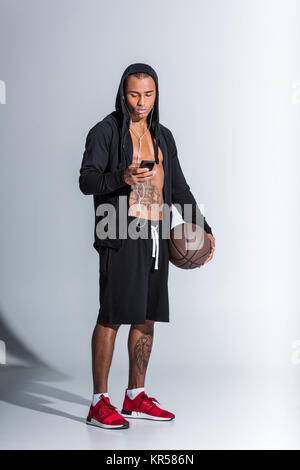 African American uomo con basket ball indossando le cuffie auricolari e utilizza lo smartphone su grigio Foto Stock