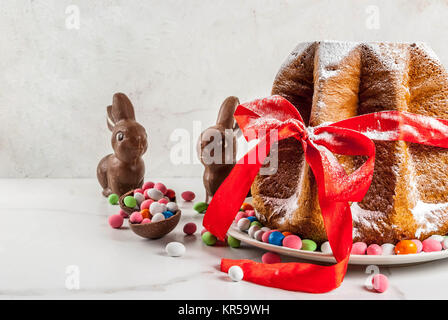 Italiano tradizionale torta di frutta Panettone Pandoro con festoso nastro rosso, Pasqua conigli e dolciumi dolce uova decorazioni, sulla casa in legno sfondo, Foto Stock