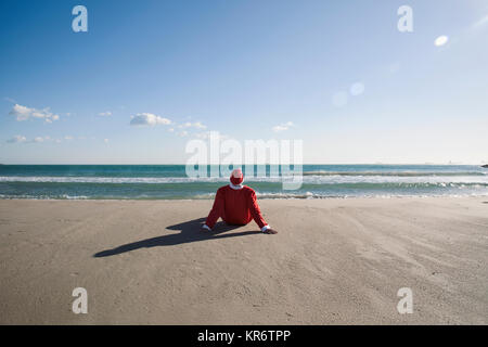 Santa Claus, visto da dietro, seduto sulla sabbia di una spiaggia rivolta verso l'orizzonte Foto Stock