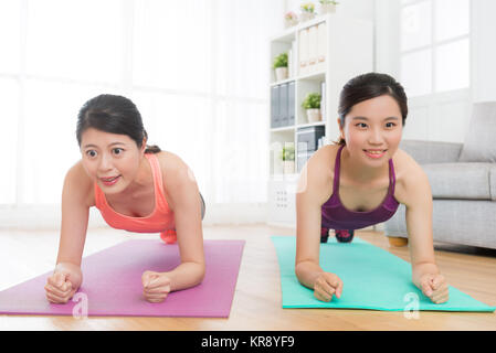 Belle ragazze sorridenti rendendo plank postura del corpo di allenamento in soggiorno e facendo un allenamento fitness muscle endurance insieme a casa. Foto Stock