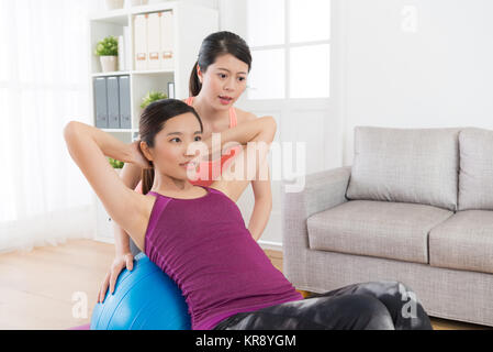 Sorridente attraente femmina studente fitness giacente sulla sfera di yoga e rendendo sgranocchia in posa nella stanza vivente con il suo allenatore per la perdita di peso. Foto Stock