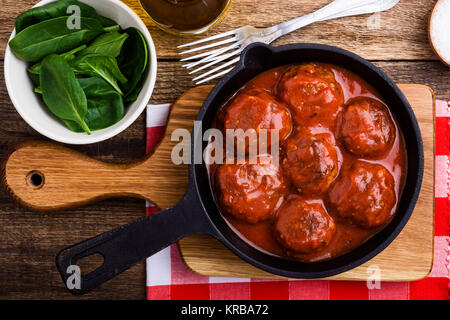 Polpette di carne in padella in ghisa con salsa di pomodoro servito fresco con insalata di spinaci su tavola in legno rustico, vista dall'alto Foto Stock