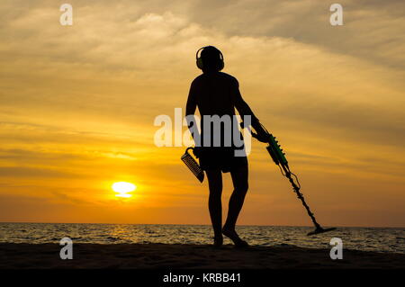 Il cacciatore di tesori con il rivelatore di metalli sul tramonto sulla spiaggia Foto Stock