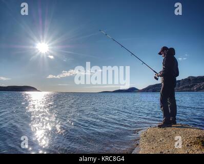 Giovane uomo la pesca sul lago al tramonto. L uomo da solo con una canna da pesca sorge sulla costa rocciosa contro il tramonto Foto Stock