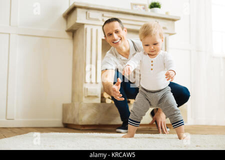 Happy baby cercando di alzarsi in piedi mentre un allegro padre essendo pronti ad aiutare Foto Stock