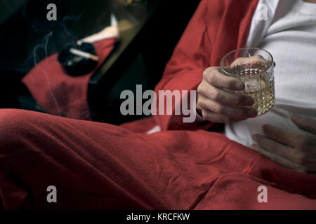 Primo piano di un giovane uomo che indossa una tuta di santa di bere whisky e fumare una sigaretta a letto Foto Stock