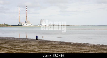 Una persona e il loro cane a camminare lungo il Sandymount Strand spiaggia sulla baia di Dublino, con il punto di riferimento twin camini di Poolbeg Power Station in aumento in la distan Foto Stock