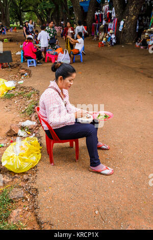 Local cambogiani donna, un venditore di souvenir a Angkor Wat, un tempio complesso vicino a Siem Reap in Cambogia, si siede sul ciglio della strada tranquillamente mangiare un pasto Foto Stock