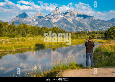 Giovani maschi adulti si erge a bordo del fiume Snake e utilizza il cellulare per scattare foto di Teton Mountain Range, Grand Tetons National Park, Teton County, Wyo Foto Stock