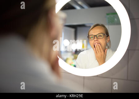 Groggy, giovane donna sbadigliare di fronte a lei lo specchio del bagno al mattino - cercando di svegliarsi e prepararsi per il lavoro (colore immagine dai toni shallow DOF) Foto Stock