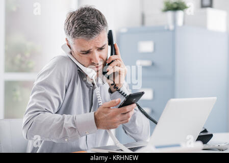 Sottolineato imprenditore disperato lavorando nel suo ufficio e avente le chiamate multiple, egli è in possesso di due telefoni e di un telefono cellulare, la gestione del business con Foto Stock