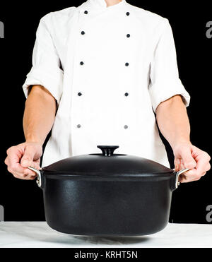 Lo Chef tenendo una casseruola verso, su tavolo bianco abbigliamento su nero Foto Stock