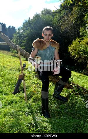 giovane arciere medievale con posta a catena seduto sul ramo, corno bevente in mano, arco e freccia accanto ad esso Foto Stock