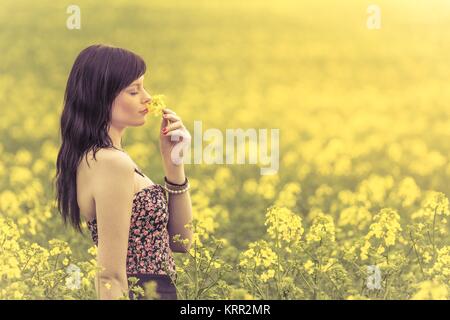 Attraktives Mädchen schnuppert un Blumen auf einer Wiese bis zum Horizont im Sonnenschein im Frühling oder Sommer. Foto einer Serie. Foto Stock