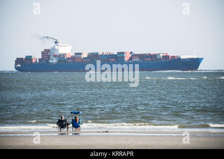 Nave container Charleston Harbor da Sullivan's ISLAND South Carolina // SULLIVAN'S ISLAND, South Carolina, Stati Uniti - Una coppia si rilassa su sedie pieghevoli sulla spiaggia all'estremità meridionale dell'isola di Sullivan mentre una grande nave container entra nel porto di Charleston sullo sfondo. Sullivan's Island, situata all'ingresso del porto di Charleston, serve sia come comunità residenziale che come popolare destinazione balneare. L'isola offre vedute del traffico marittimo che entra e lascia uno dei porti più trafficati della costa orientale. Questa punta meridionale dell'isola offre ai visitatori un'esperienza unica Foto Stock