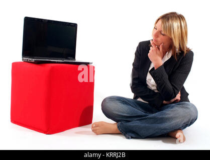 Junge Geschaeftsfrau skeptischem mit Blick sitzt neben Laptop, der auf einem roten Stoffwuerfel steht - young business donna accanto al laptop Foto Stock