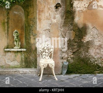 Una caratteristica e decadente angolo di un vecchio muro cadente con un vecchio ferro battuto sedia e una piccola statua Foto Stock