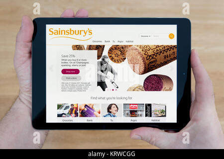 Un uomo guarda al Sainsbury's supermercato sito sul suo iPad dispositivo tablet, sparato contro un tavolo di legno top sfondo (solo uso editoriale) Foto Stock