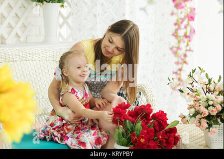 Una giovane madre con sua figlia di rilassarvi su di una terrazza estiva, che è decorato con fiori. Famiglia seduta su un rattan sedia in vimini. Foto Stock