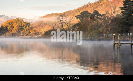 La nebbia si alza dalle tranquille acque del lago di Windermere a Ambleside, accanto a Bosco in autunno in Inghilterra del Parco Nazionale del Distretto dei Laghi. Foto Stock