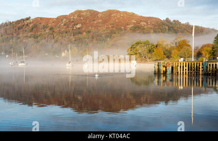 La nebbia sale dal lago di Windermere, accanto ad alberi da bosco in autunno colori, a Ambleside in Inghilterra del Parco Nazionale del Distretto dei Laghi. Foto Stock