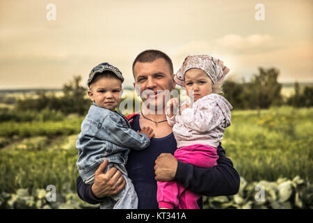 Sorridenti Padre abbracciando due graziosi bambini piccoli il figlio e la figlia in un paesaggio di simbolizzazione parenting felice Foto Stock