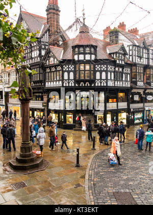 Christmas Shopper camminare su pavimenti bagnati e ciottoli nella città romana di Chester Inghilterra intorno alla croce medievale e i negozi nelle righe Foto Stock