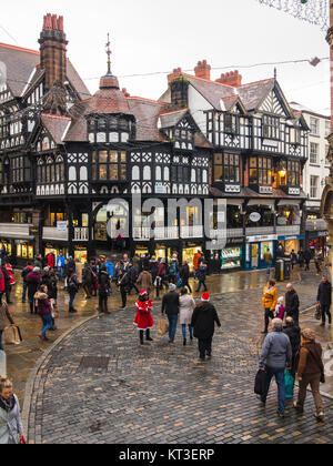 Christmas Shopper camminare su pavimenti bagnati e ciottoli nella città romana di Chester Inghilterra intorno alla croce medievale e i negozi nelle righe Foto Stock