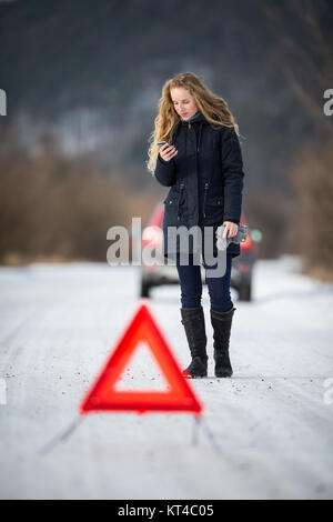 Giovane donna l'impostazione di un triangolo di avvertenza e di richiedere assistenza dopo la sua auto si è rotta nel bel mezzo del nulla su un gelido inverno giorno Foto Stock