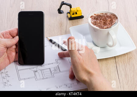 Close-up di architetto mani lavorando su un progetto e tenendo premuto uno smartphone. Tazza di caffè e estrattore in miniatura in background. Blueprint è stato creato dal fotografo. Foto Stock