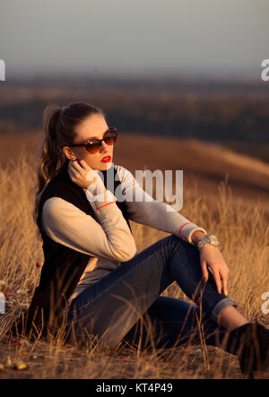 Splendida, attraente, grave ragazza con occhiali da sole seduto sulla collina con bella vista guardare il tramonto. Foto Stock