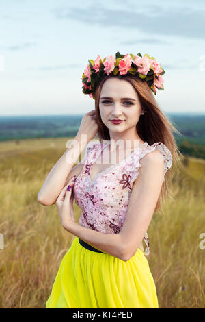 Bella piuttosto alla moda splendida ragazza in abiti sul campo dei fiori. Bella ragazza con la corona di fiori sul suo capo e bouquet di molti fiori gialli seduta sul prato campo giallo in estate. Foto Stock