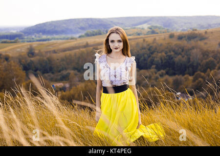 Bella piuttosto alla moda splendida ragazza in abiti sul campo dei fiori. Bella ragazza con la corona di fiori sul suo capo e bouquet di molti fiori gialli seduta sul prato campo giallo in estate. Foto Stock