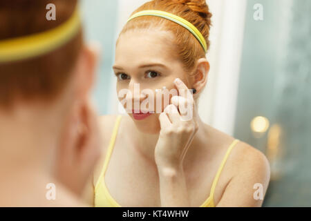 Redhead girl l'applicazione di crema di bellezza nel bagno di casa al mattino. Giovane donna prendersi cura della sua pelle, guardando a specchio in slowmotion. Bellezza femminile, s Foto Stock