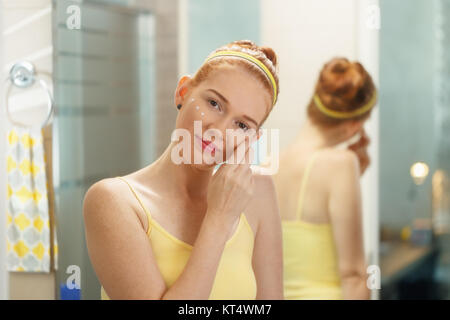 Redhead girl l'applicazione di crema di bellezza nel bagno di casa al mattino. Giovane donna prendersi cura della sua pelle, cercando intimamente alla fotocamera. Bellezza femminile, pelle Foto Stock
