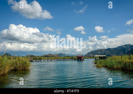 Un capanno sulla estremità di un molo di legno in una distanza su un lago con lamelle di verde sotto un cielo azzurro con soffici nuvole bianche Foto Stock